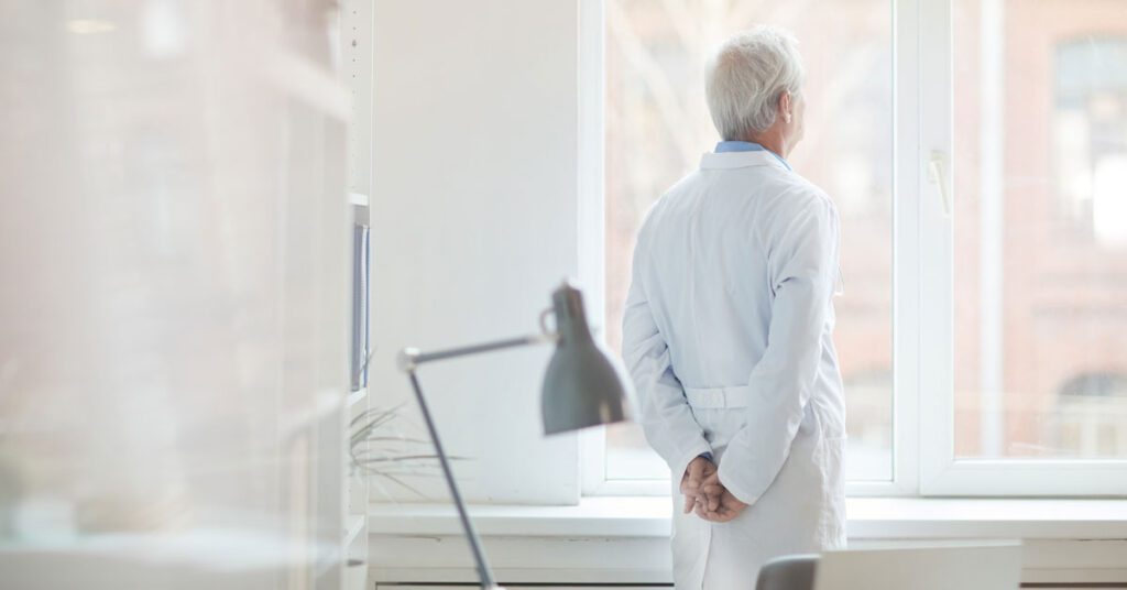 Senior doctor standing with hands behind his back, looking out of a window
