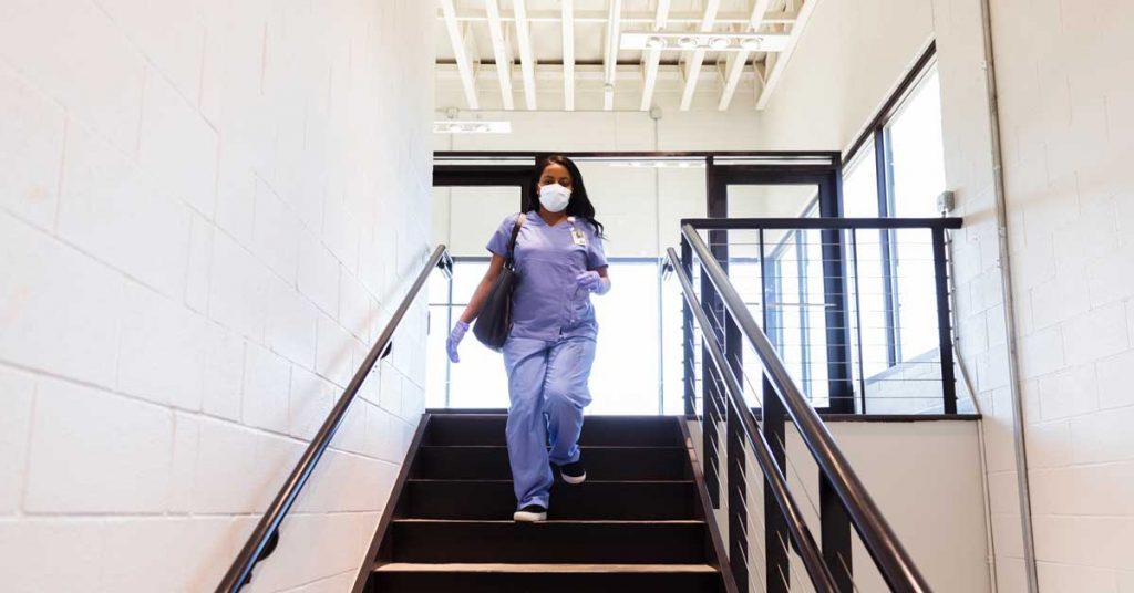 Healthcare worker wearing scrubs and a mask, walking down a stairwell in a bright, modern building