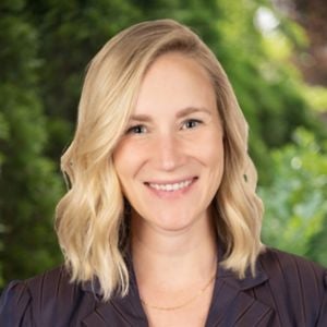 Helen Falkner smiling with shoulder-length blonde hair wearing a dark blazer in front of a green foliage background