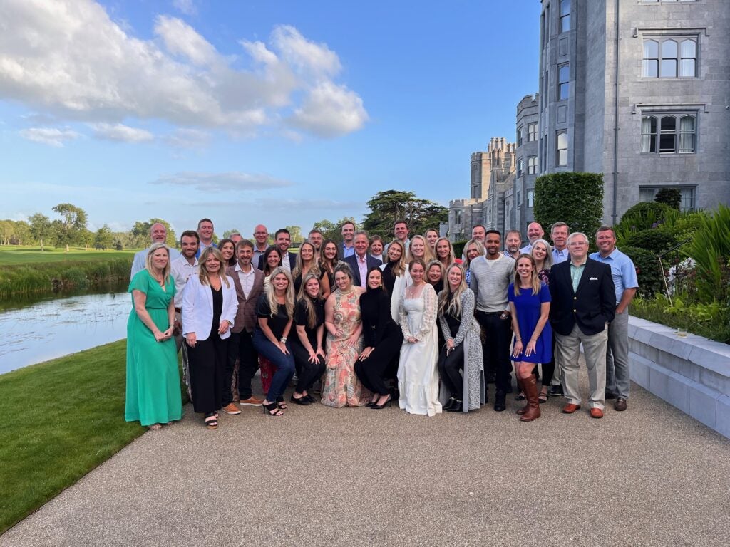 A large group of people posing together outside a historic building, enjoying a sunny day with clear blue skies and lush greenery in the background
