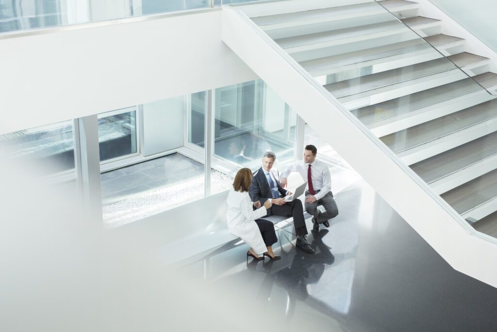 Three professionals having a discussion while seated on a bench under a large, modern staircase