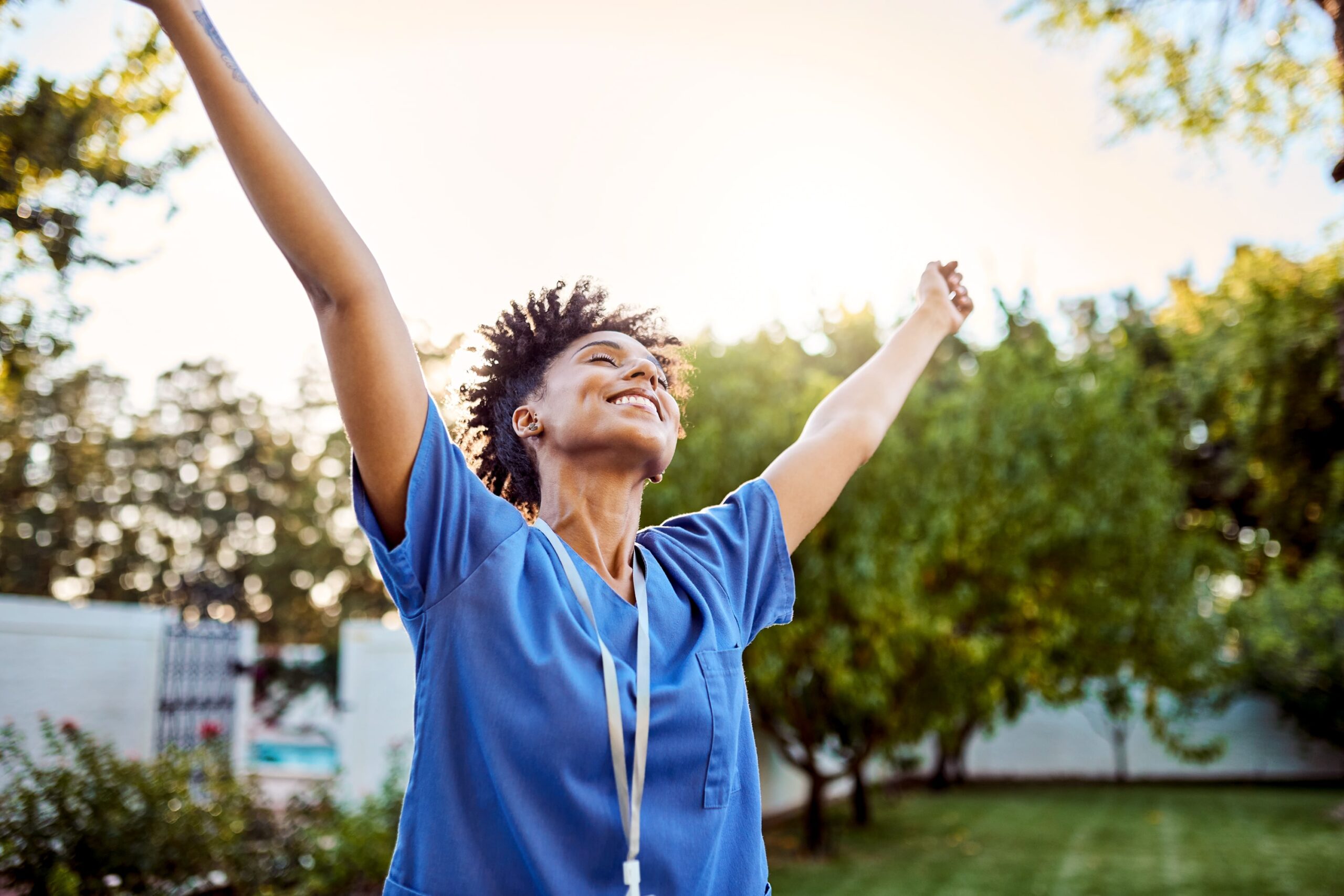 young woman smiling in blue scrubs with her arms outstretched to the sky