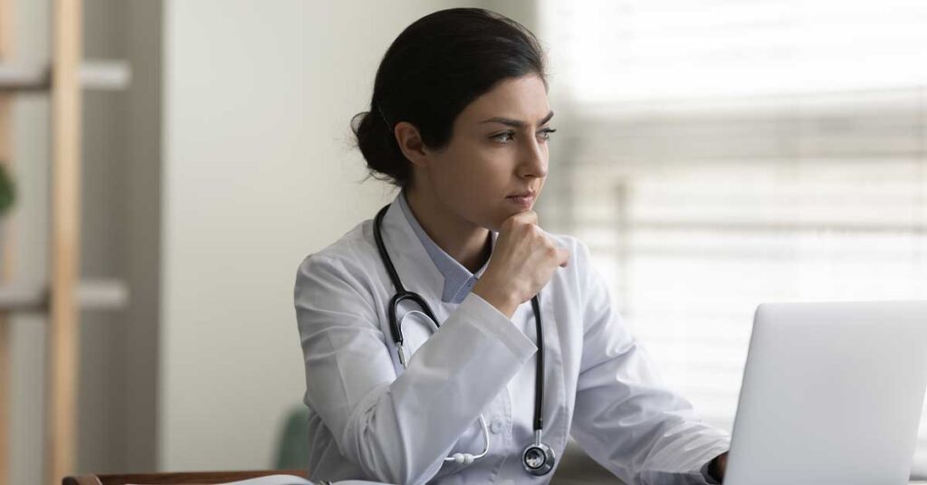 A female doctor in a white coat sitting at a desk, looking thoughtfully at a laptop