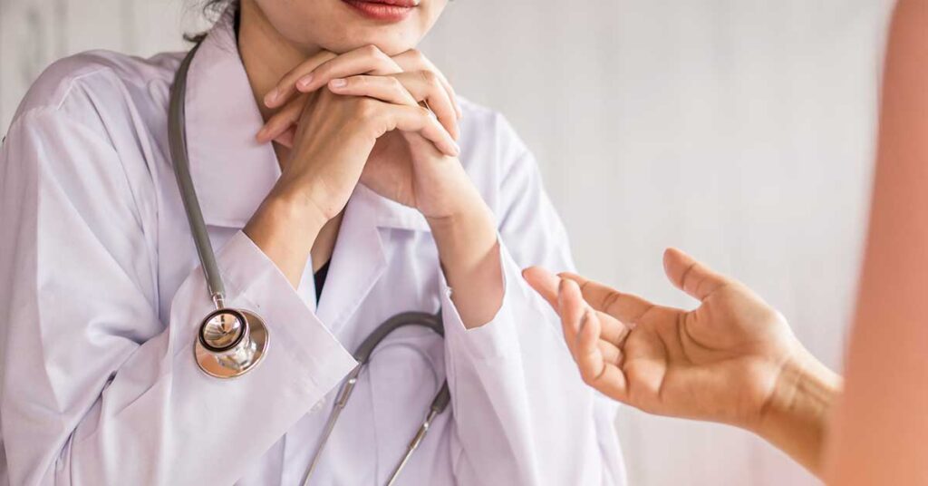 A close-up of a doctor in a white coat with a stethoscope around their neck, listening attentively to a patient whose hand is gesturing while speaking