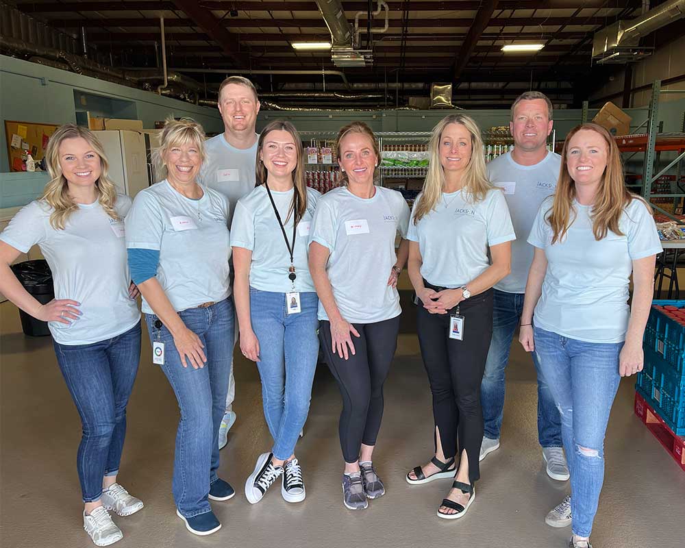 A group of eight people wearing light blue shirts standing together in a warehouse, smiling at the camera