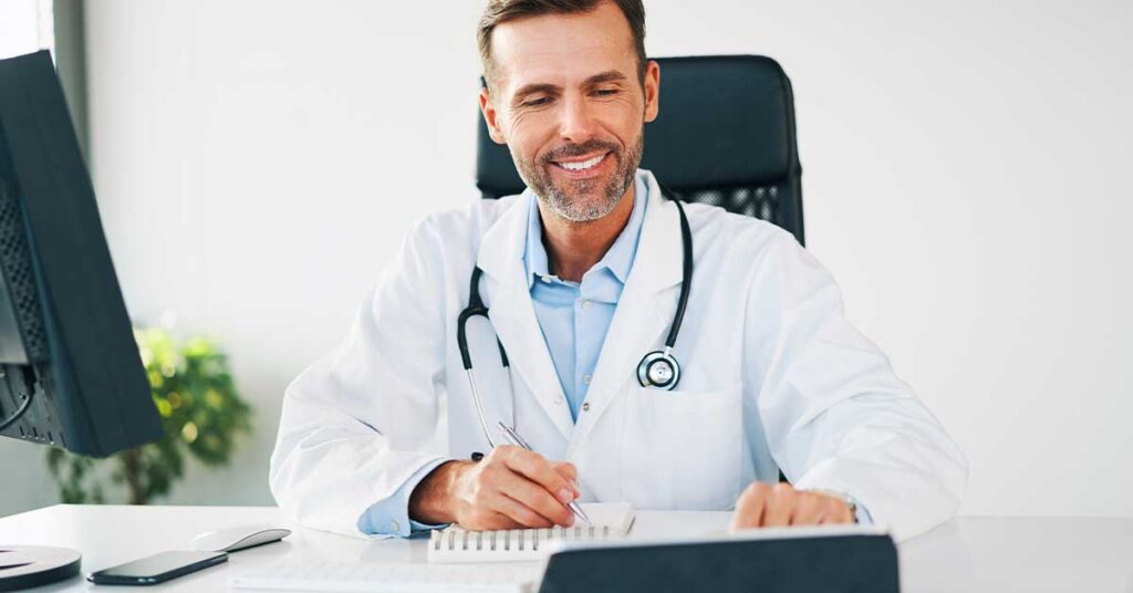 Smiling male doctor in a white coat with a stethoscope, writing at a desk