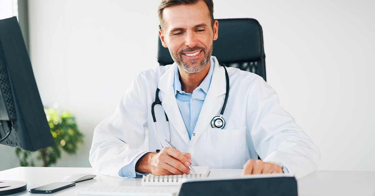 Smiling male doctor in a white coat with a stethoscope, writing at a desk