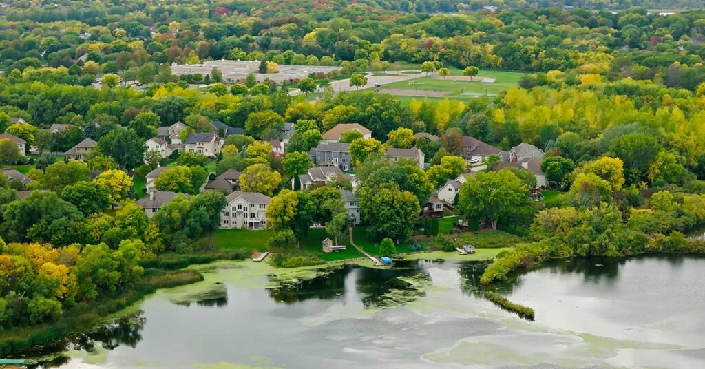 Aerial view of a suburban neighborhood with green trees, a lake in the foreground, and houses of varied architectural styles