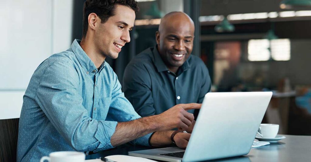 Two men in a modern office smiling while discussing something on a laptop, with coffee cups on the table
