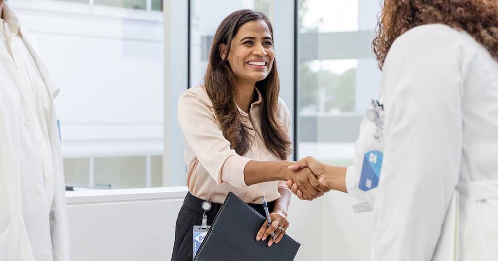 Smiling professional in a business setting shaking hands with a healthcare worker, symbolizing partnership in healthcare