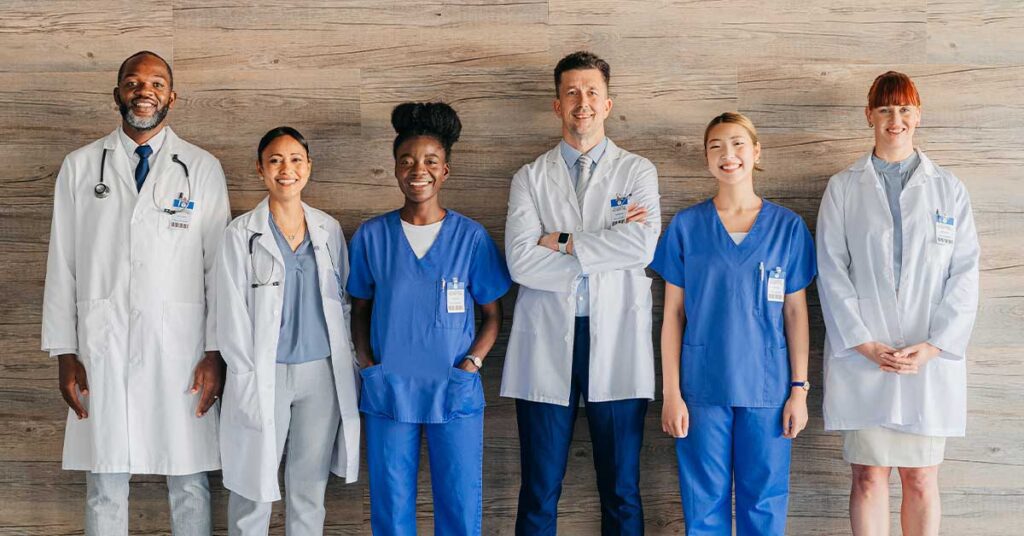 Diverse healthcare staff, including doctors and nurses, standing confidently together in front of a wooden wall backdrop