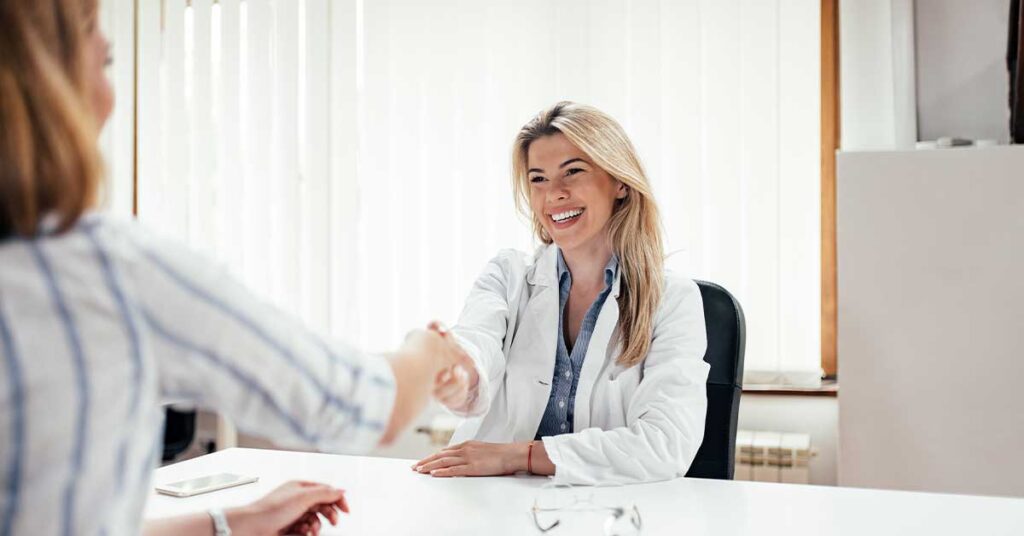 Smiling female doctor in a white coat shaking hands with a patient in a medical office