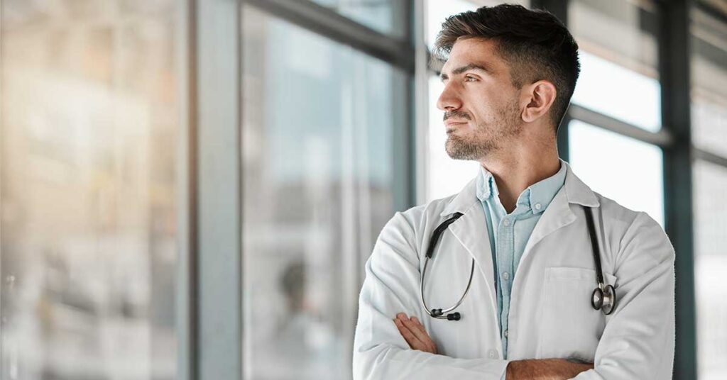 Thoughtful male doctor in a white coat with a stethoscope, standing by a hospital window