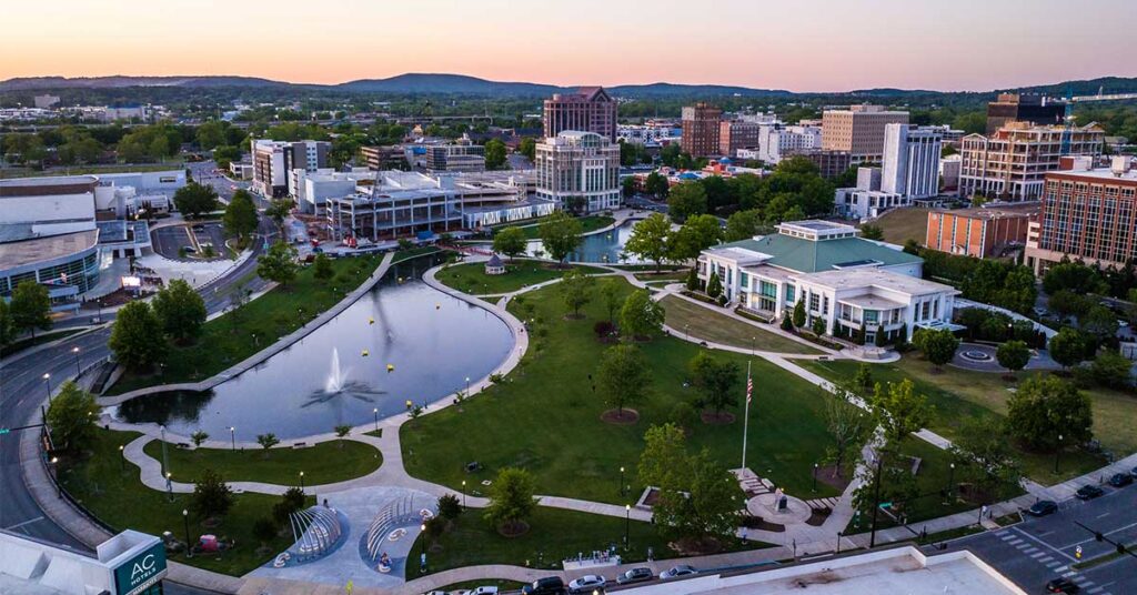 Aerial view of a modern city park with a large pond, walking paths, and surrounding urban buildings at dusk