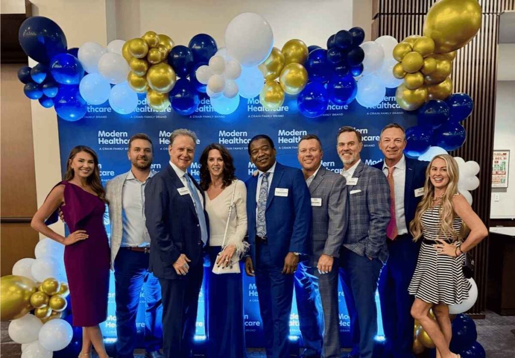 Group of professionals posing together at a "Modern Healthcare" event with a backdrop of blue, white, and gold balloons.
