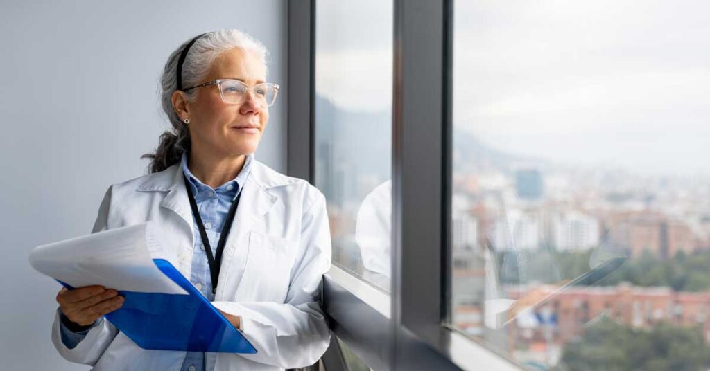 Latin American female doctor holding a clipboard at the hospital and smiling while looking through the window