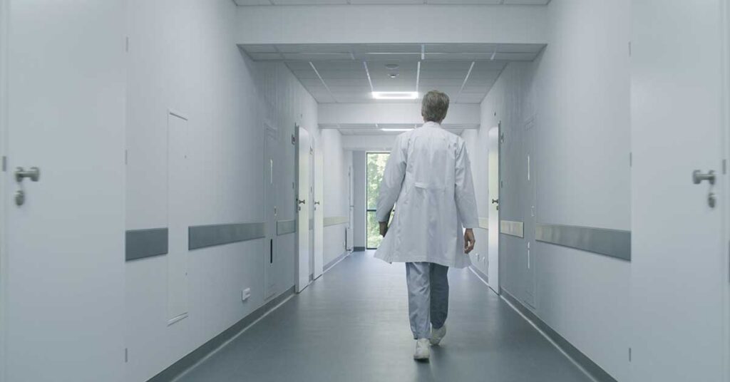 Mature doctor walks down hospital corridor with digital tablet computer to medical room.