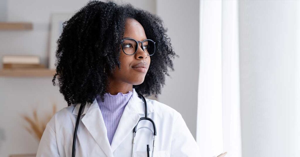 Professional african-american female doctor wearing glasses holds a clipboard while thoughtfully standing in a bright medical office space