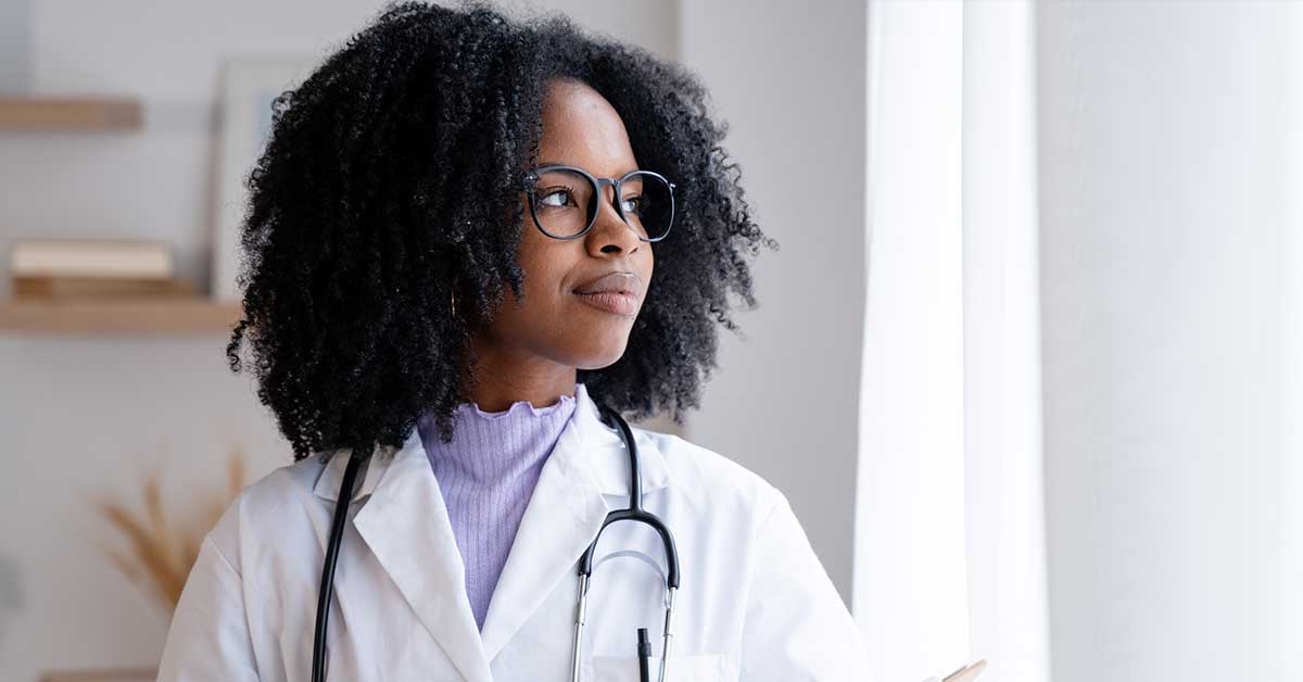 Professional african-american female doctor wearing glasses holds a clipboard while thoughtfully standing in a bright medical office space