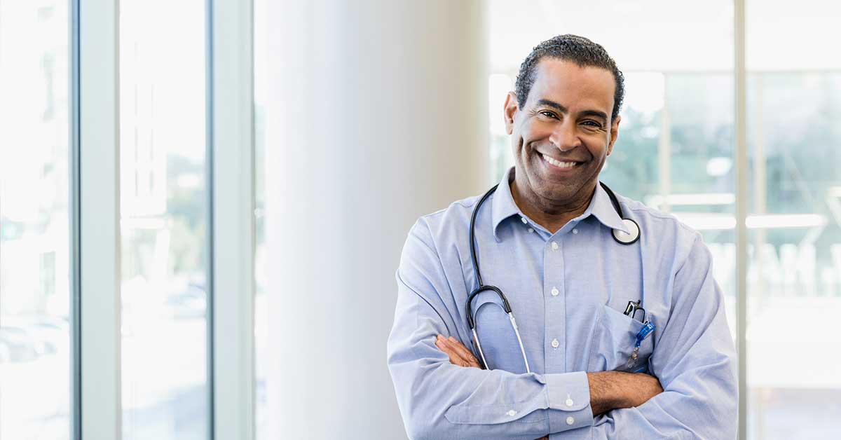 Confident mature male doctor stands with crossed arms in office