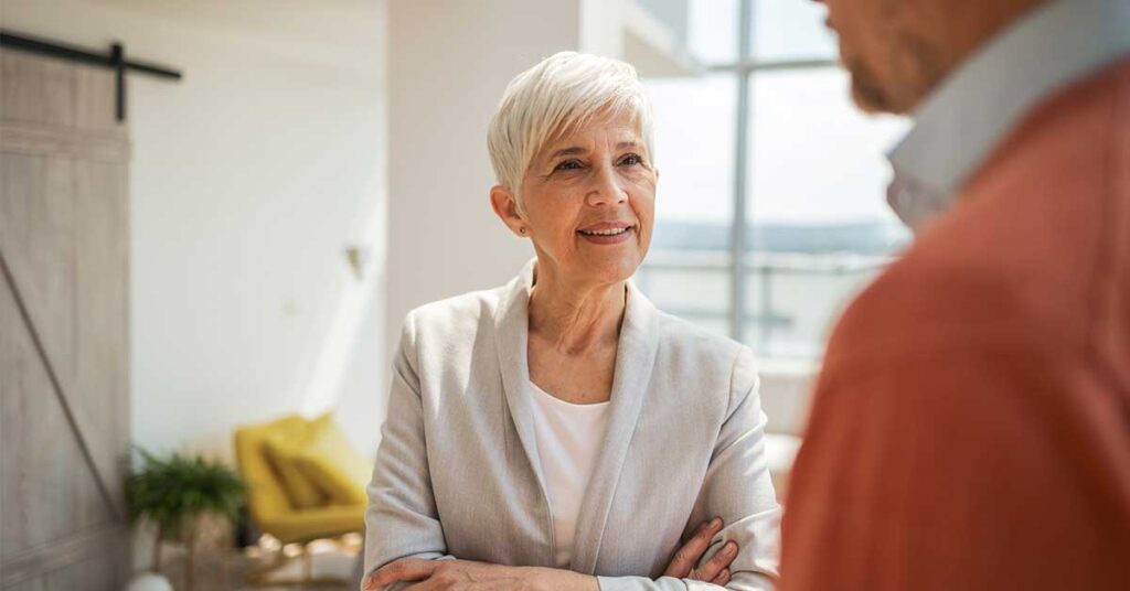 Elderly professional woman standing thoughtfully, speaking to a colleague in a spacious, brightly lit office.