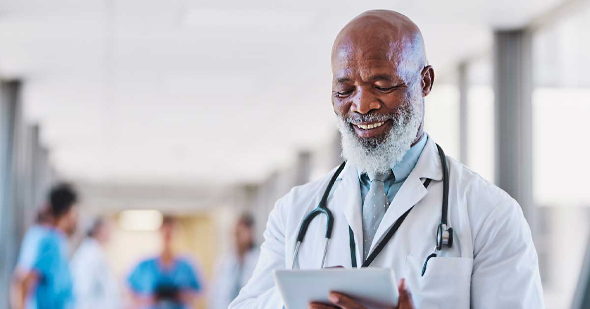 Senior specialist with beard using and smiling cheerfully at digital tablet in hospital corridor, coworkers in background