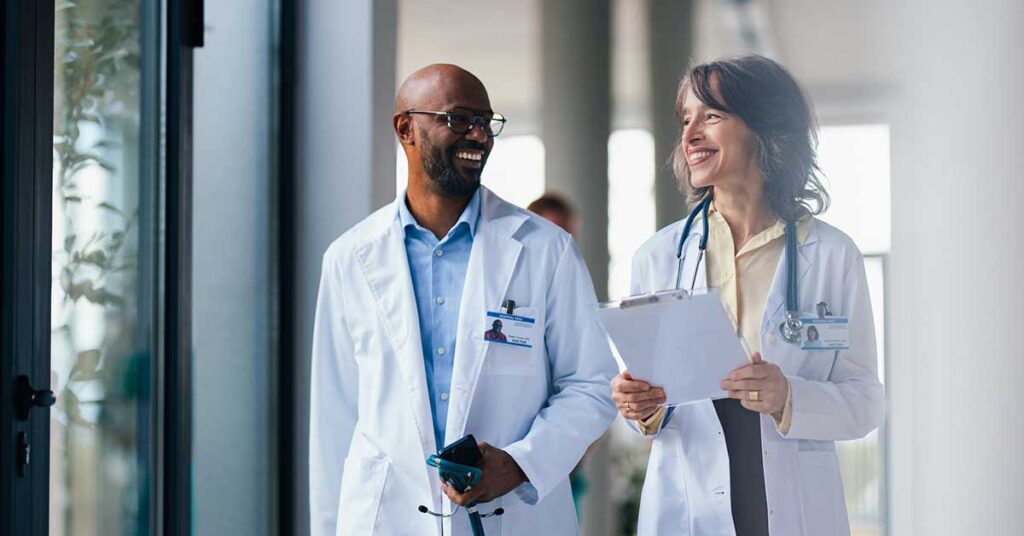 wo doctors in white coats share a light moment while walking through a hospital corridor