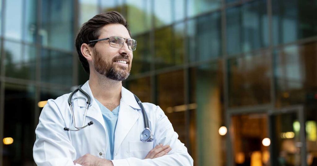 Confident doctor wearing a white lab coat and stethoscope, standing with arms crossed and smiling while looking away from a modern hospital building facade