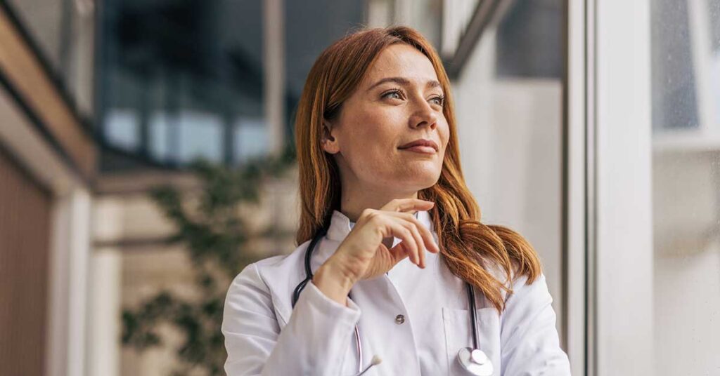 Thoughtful female doctor in a lab coat and stethoscope gazing out the window, reflecting on her responsibilities and the future of healthcare in the hospital setting